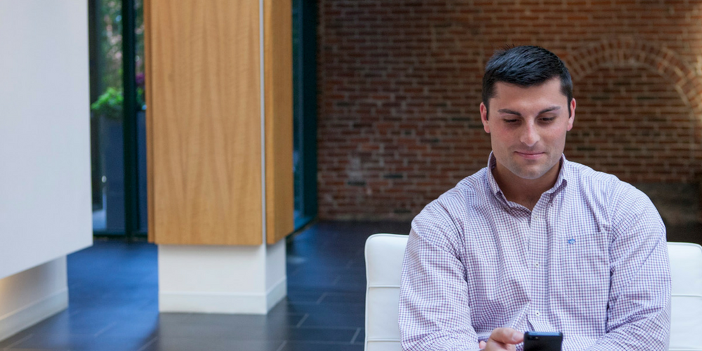 A man in a light checkered shirt sits on a white chair indoors, looking down and smiling slightly at his phone. The background includes a brick wall and modern architectural elements.