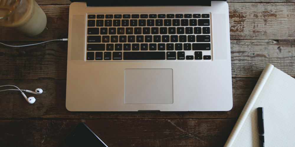 A silver laptop on a rustic wooden table, surrounded by white earphones, a smartphone, a notebook, and a glass of iced coffee—perfect for staying productive while managing your real estate business.
