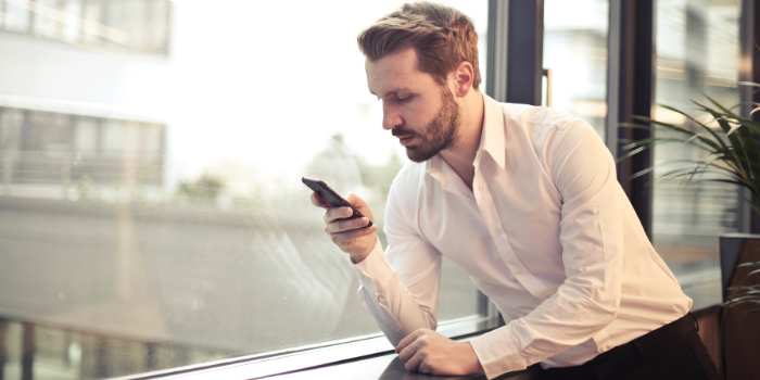 A man in a white shirt leans against a windowsill, looking intently at his smartphone. He is indoors, near large windows with natural light and a potted plant visible beside him.