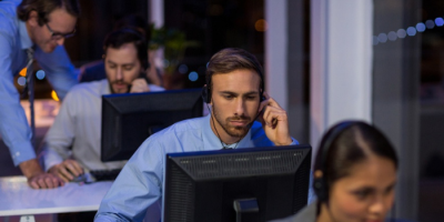 Several people wearing headsets sit at desks with computers in an office at night. One man in the foreground is focused on his screen, while another man stands behind him, assisting a colleague.