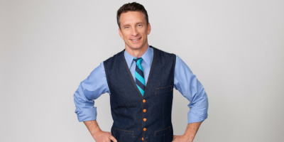 A smiling man with short brown hair stands with hands on hips, wearing a light blue shirt, a blue striped tie, and a dark denim vest with brown buttons, against a plain light background.