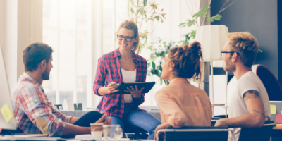 Four young adults sit and talk in a bright, modern office with plants. One woman stands, smiling and holding a tablet, while the others discuss ideas about Live Receptionist Services.