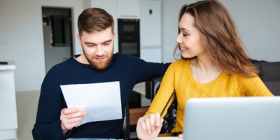 5-Things-Accountants-Need-To-Do-Before-Next-Tax-Season A man and woman sit together at a table, looking at a paper the man is holding. The woman smiles at him while using a laptop. They appear to be in a bright, modern home.