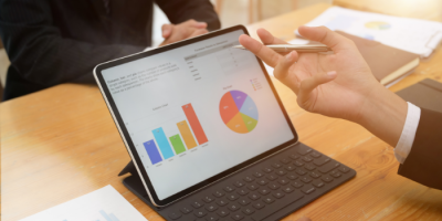 Two people in business attire sit at a wooden table with a tablet displaying colorful charts and graphs. One, the Abby Connect CEO, gestures toward the screen with a pen, engaging in an insightful discussion about the data.