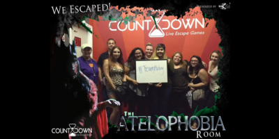 A group of ten people smile and pose together, holding a sign that reads #TeamFabtay and Abby Connect in front of a red wall at Countdown Live Escape Games. Text around the image says We Escaped! and The Atelophobia Room.