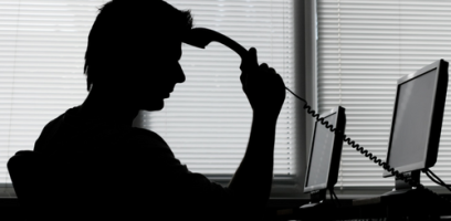 Silhouette of a person sitting at a desk with two monitors, holding a corded phone handset to their forehead, appearing stressed or contemplative, with blinds covering the window in the background.