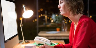 A woman in a red blouse sits at a desk, working on a computer at night. A desk lamp, phone, papers, and office supplies are visible, with blurred lights in the background.