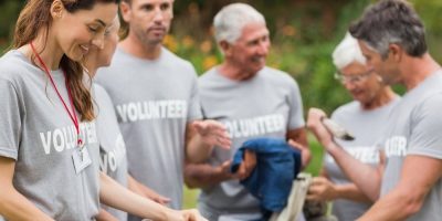 A group of smiling people wearing matching grey Volunteer shirts work together outdoors, engaging in a community service activity. Some hold tools and bags, suggesting they are cleaning or collecting items.