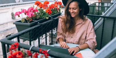 A woman wearing a hat and pink shirt smiles while working on a laptop at a small balcony table, surrounded by blooming red and pink flowers in planter boxes.