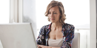 A woman with short, wavy hair wearing a patterned shirt sits at a desk, working on a laptop near a bright window.