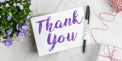 A card with the words Thank You in purple script sits open on a marble surface beside a black pen, a small potted plant with purple flowers, and a white gift box wrapped with red and white twine.