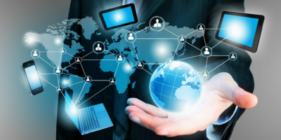 A person in a suit holds a glowing digital globe, with icons of laptops, tablets, and smartphones connected by lines overlaying a world map, symbolizing global digital connectivity and technology.