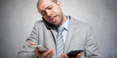 Why-Overflow-Phone-Answering-Helps-Your-In-House-Receptionist-Do-More-1 A man in a gray suit and striped tie is talking on one phone while holding two other phones, looking stressed and overwhelmed, against a plain gray background.