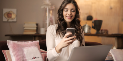 A woman with long brown hair sits on a couch, smiling while looking at her smartphone. She has a laptop on her lap and is surrounded by pillows and books in a cozy, softly lit room.