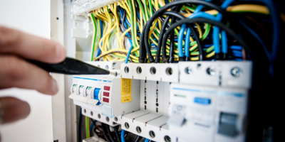 A hand uses a tool to adjust or inspect a circuit breaker in an electrical panel filled with colored wires and switches.