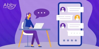 Illustration of a woman with a headset working on a laptop at a desk, with a large smartphone screen showing chat messages and star ratings. The Abby Connect logo is in the top left corner on a purple background.