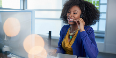 Smiling woman wearing a headset sits at a desk in an office, looking at a computer screen while typing, suggesting she is providing Call Answering Services or working in customer support.
