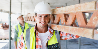 Two construction workers wearing white hard hats and safety vests carry a wooden beam at a building site. One worker smiles at the camera while the other is in the background, slightly out of focus.