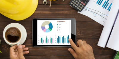 A person uses a tablet displaying charts and graphs at a desk with a cup of coffee, a yellow hard hat, calculator, rolled papers, and printed charts.