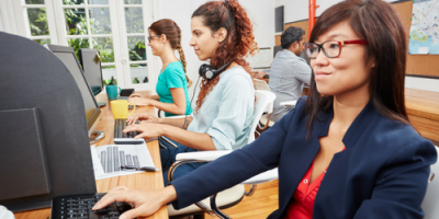 Four people working at desks in a modern office, focused on their computer screens. The workspace has large windows, plants, and a corkboard on the wall. One person has headphones around their neck.