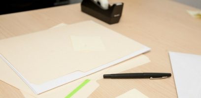 A desk with a manila folder, a black pen, a green highlighter, loose papers, a sticky note, and a tape dispenser.
