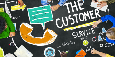 Several people gather around a table covered with drawings and notes about customer service, including a large phone receiver, chat bubble, and the words THE CUSTOMER and SERVICE written prominently.