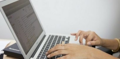 Close-up of a person’s hands typing on a silver laptop keyboard, with an email inbox open on the screen. A closed Lenovo notebook lies next to the laptop on the desk.
