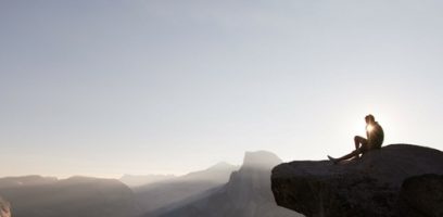 customer_service_blog_15-11 A person sits at the edge of a rocky cliff, silhouetted against the sky at sunrise or sunset, overlooking distant mountains and a hazy landscape.
