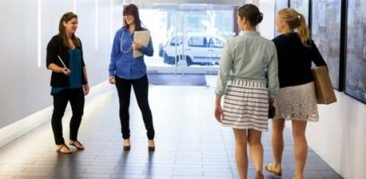 Four women are walking in a bright hallway. Two are talking on the left, one holding a notebook, while two others walk away from the camera on the right, carrying bags, dressed in casual clothing. A car is visible outside through glass doors.