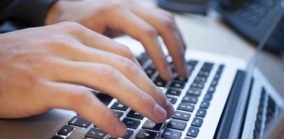 Close-up of a persons hands typing on a laptop keyboard. The image focuses on the fingers pressing the keys, with the laptop and part of the desk visible in the background.