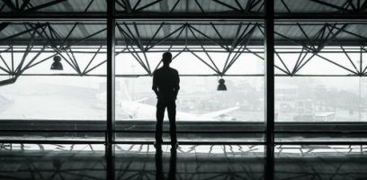 customer_service_blog_18 A person stands alone, silhouetted against large windows at an airport terminal, perhaps awaiting a flight after using a Business Answering Service, gazing out at a plane on the tarmac on a foggy day.