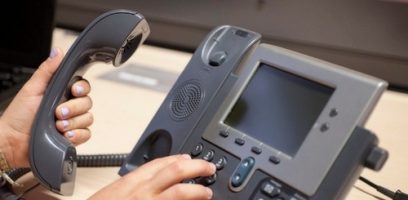 customer_service_blog_2-101 A person with light purple nail polish holds a handset and dials numbers on a gray office desk phone with a screen, buttons, and a coiled cord.