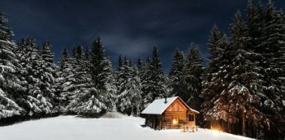 A small wooden cabin with glowing lights sits among snow-covered pine trees under a starry night sky in a winter forest.