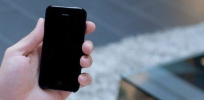 A hand is holding a black smartphone with the screen turned off. The background is slightly blurred, showing a gray floor and a textured, light-colored surface.