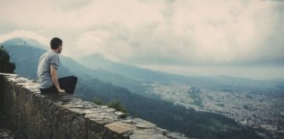 A person sits on a stone wall overlooking a city nestled among mountains, with a cloudy sky above and mist rolling over the distant hills.