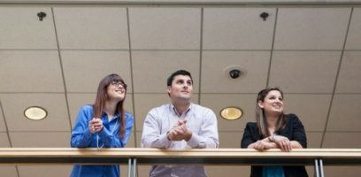 customer_service_blog_25-11 Three people stand indoors at a railing, looking ahead and smiling. The ceiling with lights and a security camera is visible above them.