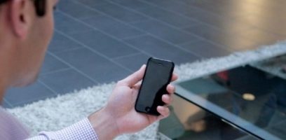 customer_service_blog_26-1 A person holding a black smartphone with a blank screen, sitting near a glass table and a light-colored rug on a tiled floor.