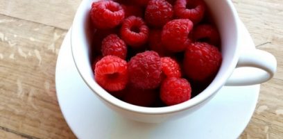 A white cup filled with fresh, bright red raspberries sits on a matching white saucer on a wooden surface.