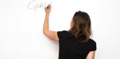 A woman with brown hair, seen from behind, writes the word GOALS on a whiteboard with a black marker.