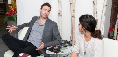 Two people sit and talk in a modern office lounge; the man gestures while speaking, and the woman listens. A small table with magazines and keys is in front of them, and there are birch branch decor and plants behind them.