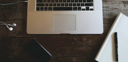 A laptop on a wooden desk with earphones, a smartphone, an open notebook, and a pen arranged around it.