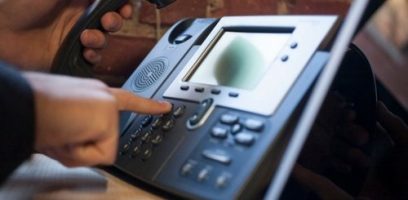 A person is dialing a number on a black office desk phone while holding the handset, with a brick wall in the background.