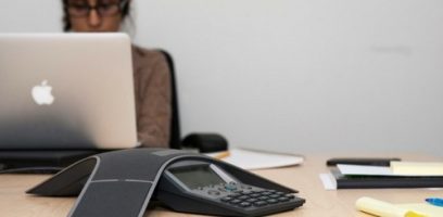 A person sits at a desk working on a laptop with an Apple logo. In the foreground, there is a conference speakerphone and some office supplies on the table.