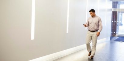 A man in business casual attire walks down a bright hallway while looking at his smartphone. Vertical light panels are mounted on the white wall beside him.