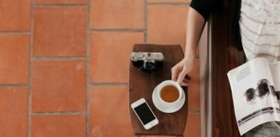 A person holds a cup of coffee at a wooden table with a camera, smartphone, and an open magazine on it, next to terracotta floor tiles.