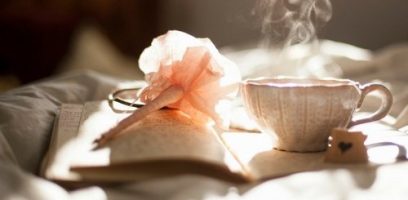 A steaming cup of tea sits on an open notebook with a pink, flower-shaped pen resting beside it, bathed in soft, warm sunlight.