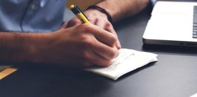 A person writes on a piece of paper with a yellow pen next to an open laptop on a dark desk.