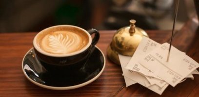 customer_service_blog_53 A cup of latte with leaf-shaped foam art sits on a saucer next to a stack of small receipts and a brass service bell on a wooden table.
