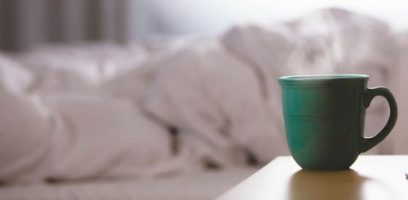 customer_service_blog_57 A green ceramic mug sits on a bedside table in the foreground, with an unmade bed and soft rumpled white bedding blurred in the background.