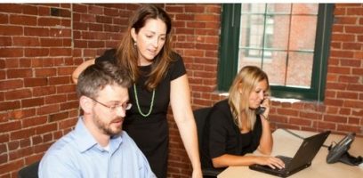 Three people work in an office with brick walls; a man uses a computer, a woman stands beside him looking at the screen, and another woman speaks on the phone while using a laptop.
